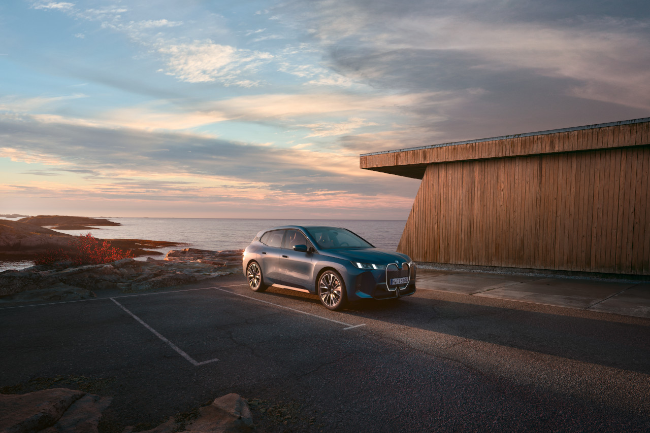 Dark blue BMW SUV parked on an empty asphalt lot by the coast at sunset, next to a modern wooden building.
