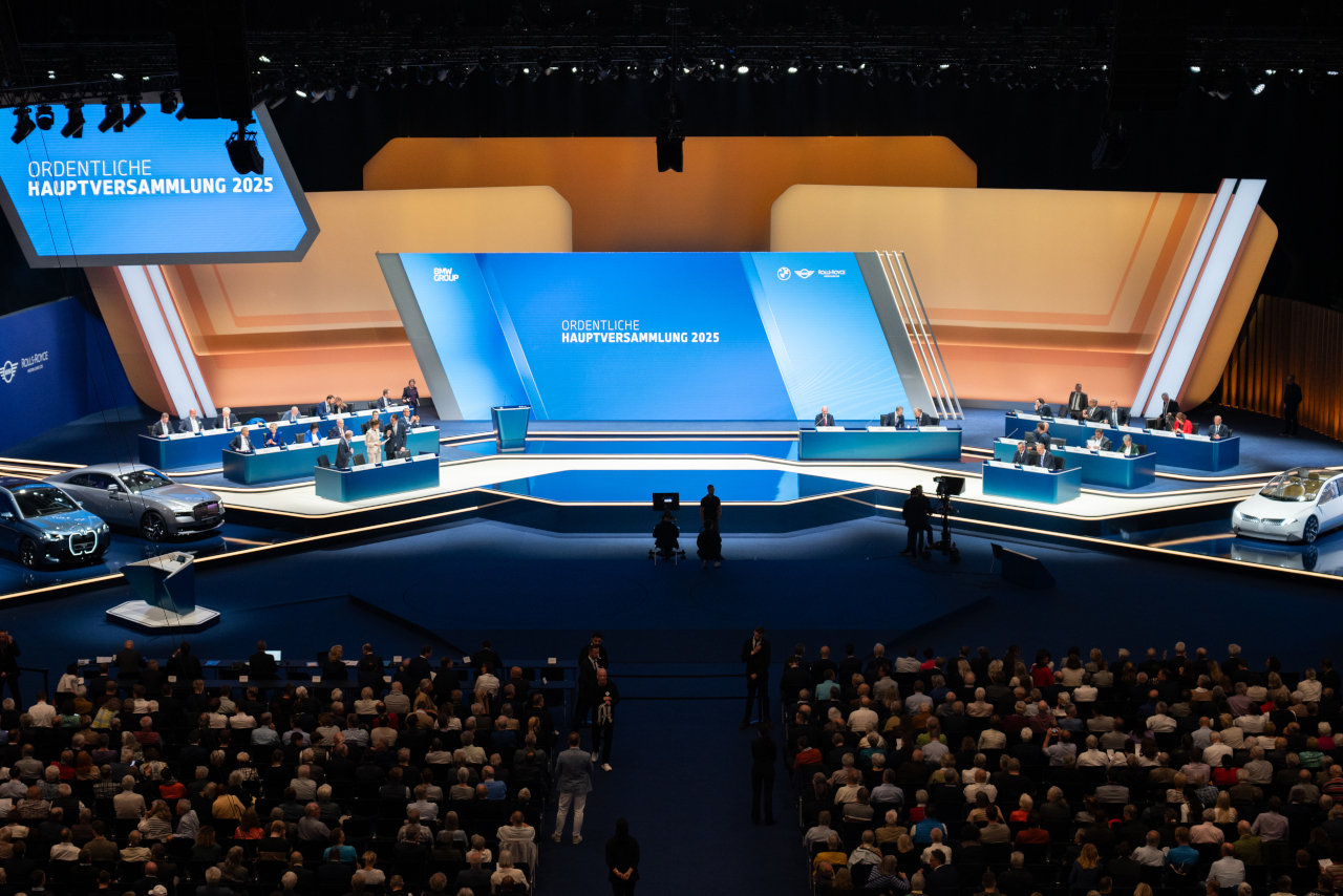 BMW Annual General Meeting 2025 in Munich: Large-scale event with stage, lecterns, displayed vehicles, and a large audience.