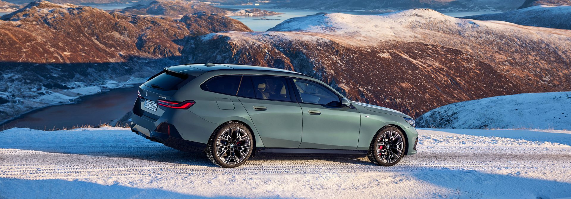 A BMW i5 Touring in metallic green on the road in a snowy mountain landscape. 