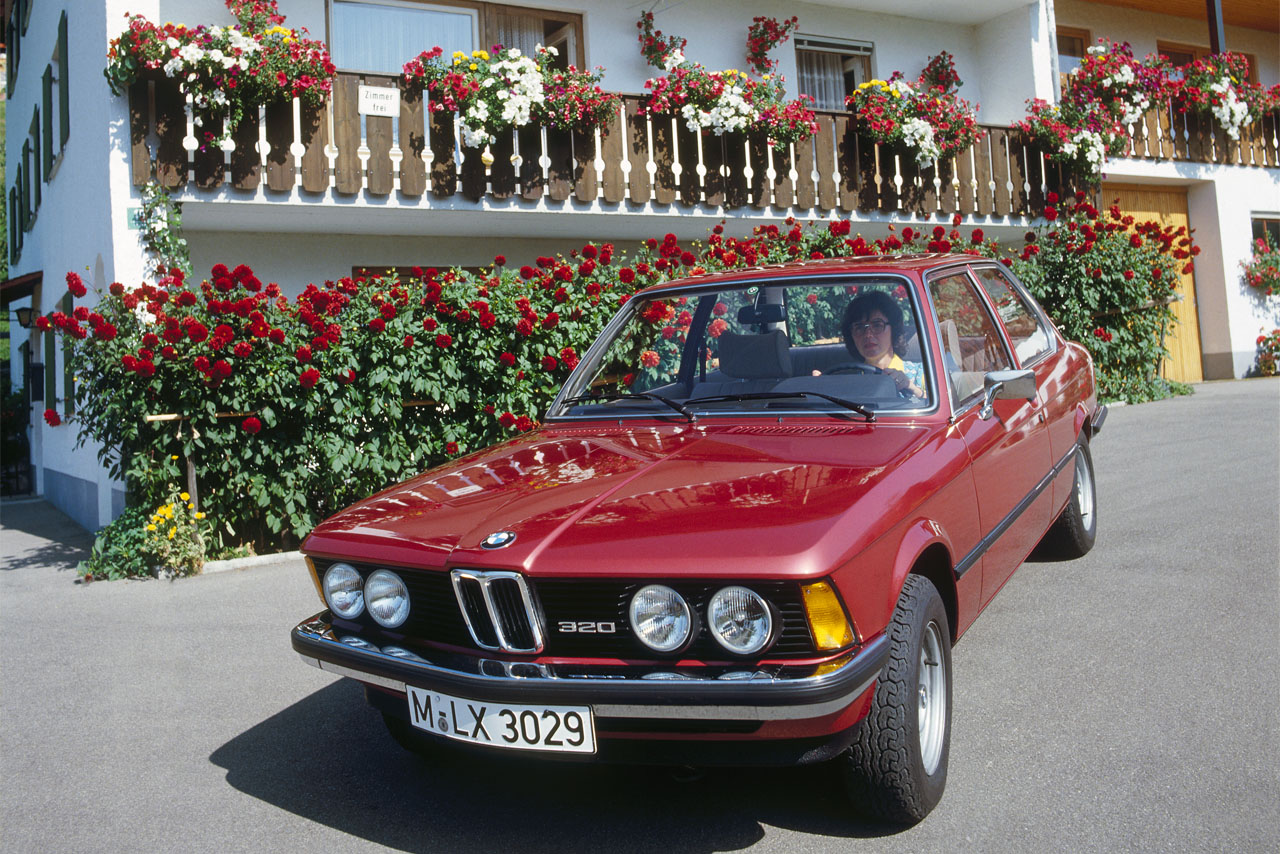 A red BMW 320 (E21) with double headlights in front of an alpine building with flowers on the balconies.