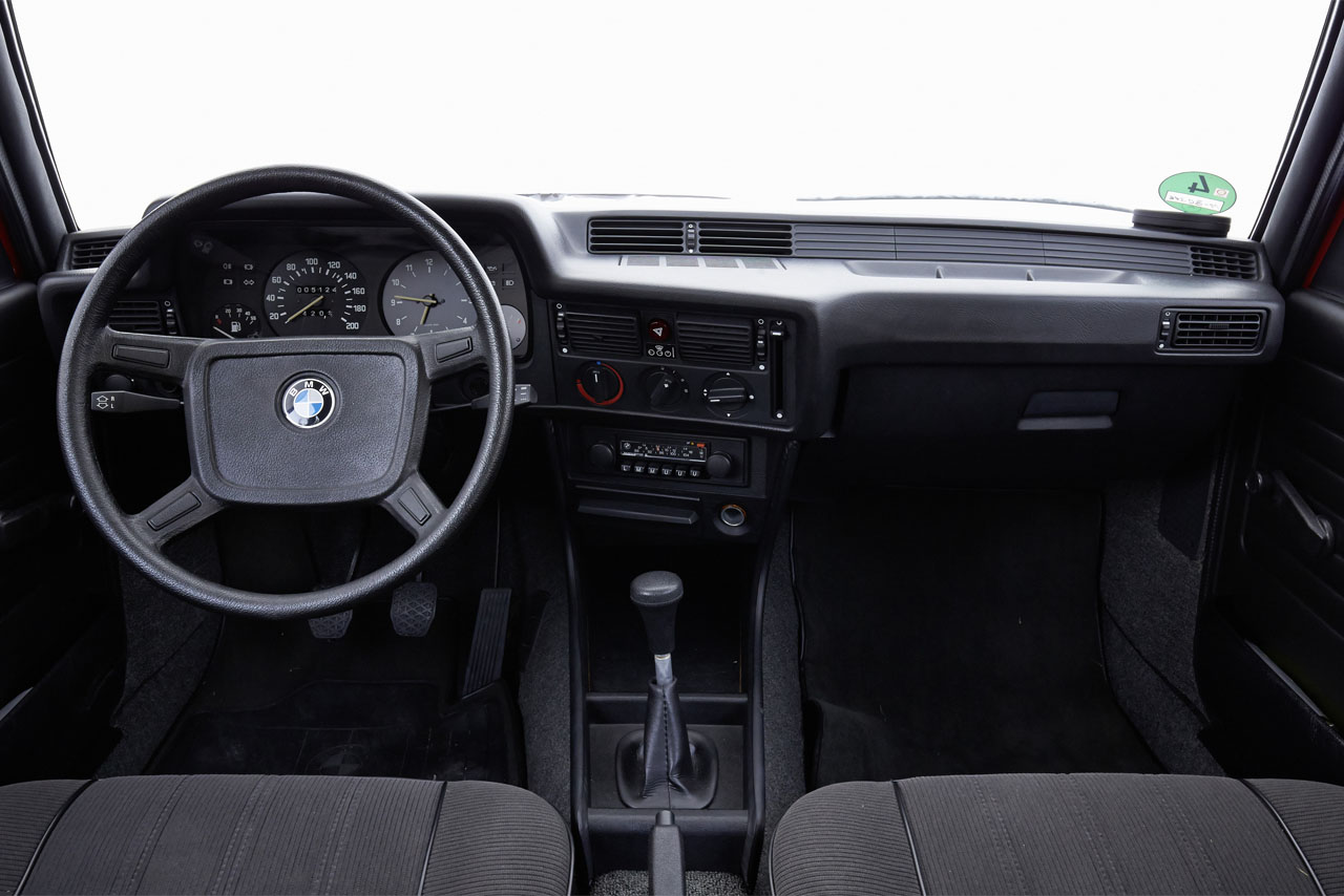 The classic BMW E21 interior with a cockpit in black: The steering wheel, analogue dials, centre console and clear dashboard layout, seen from the rear seat.