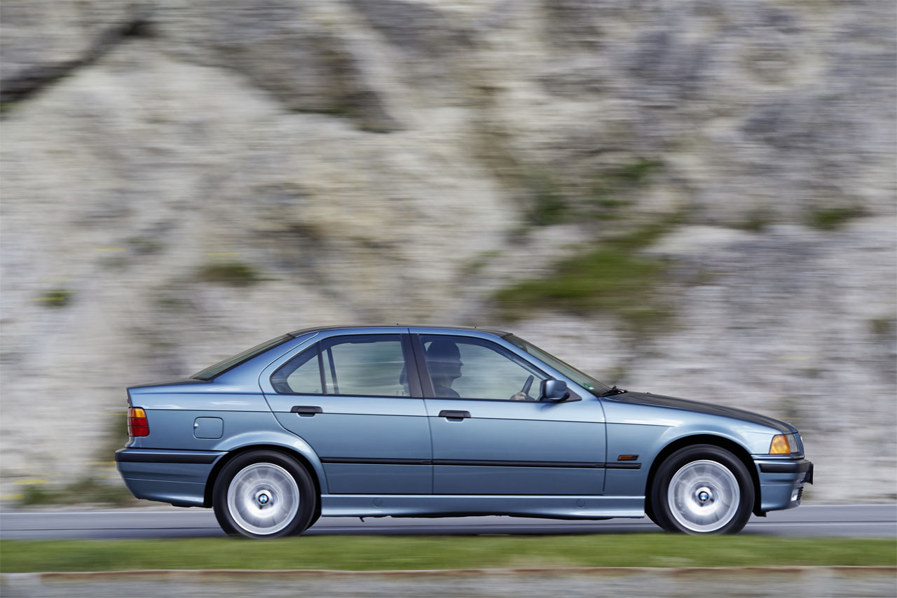A silver-blue BMW E36 Sedan seen from the side as it drives dynamically past a rockface.