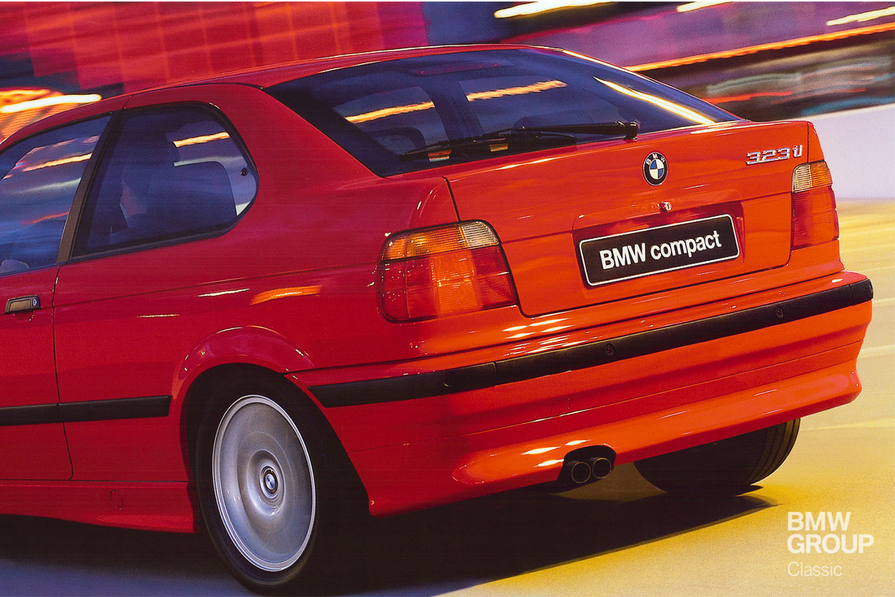 A red BMW 323ti in motion: View of its short rear and left side, with dynamic lighting in the background.