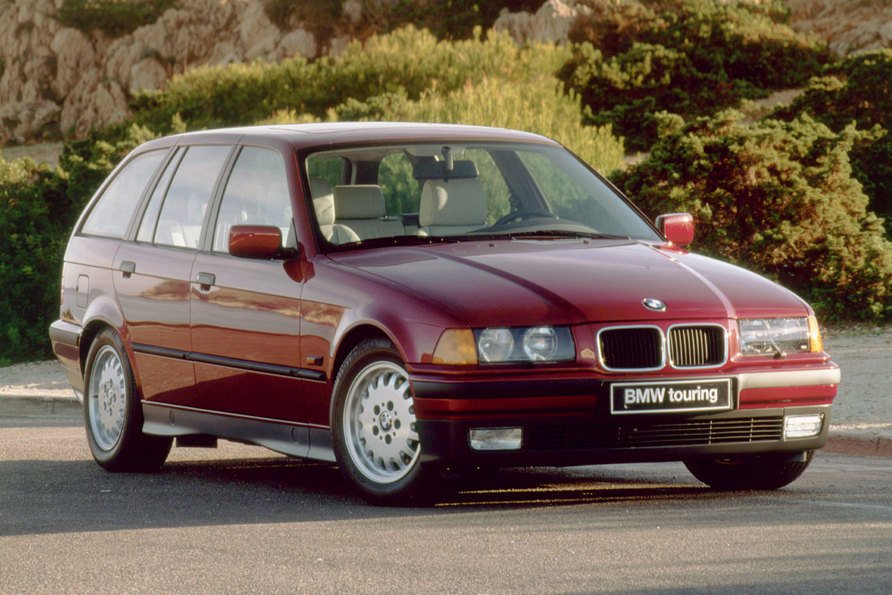 A dark red BMW E36 Touring seen from the front right with lush, green vegetation in the background.