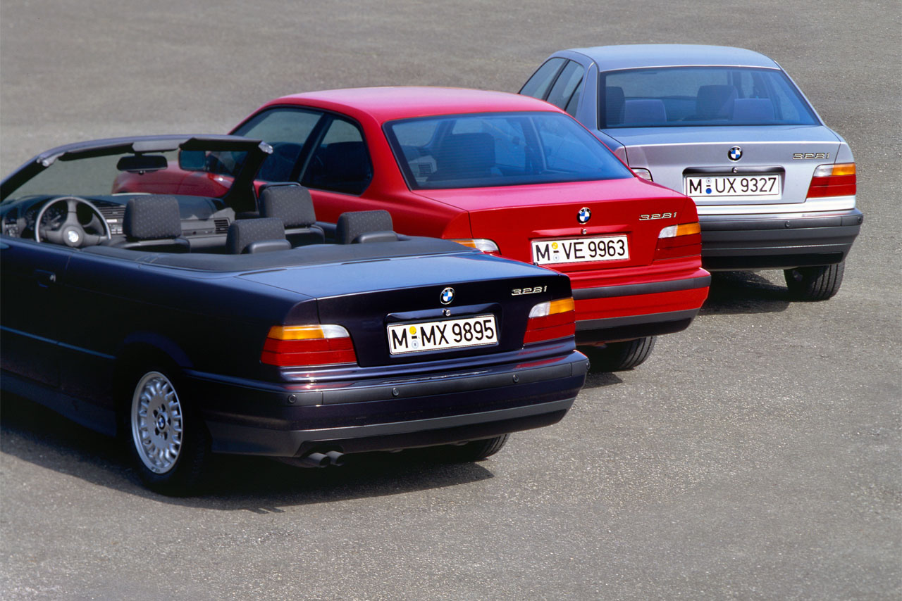 Three BMW E36 models seen from the rear: An open-topped Convertible in dark blue (front), a Coupe in red (centre), and a Sedan in silver (back), arranged diagonally in a row.