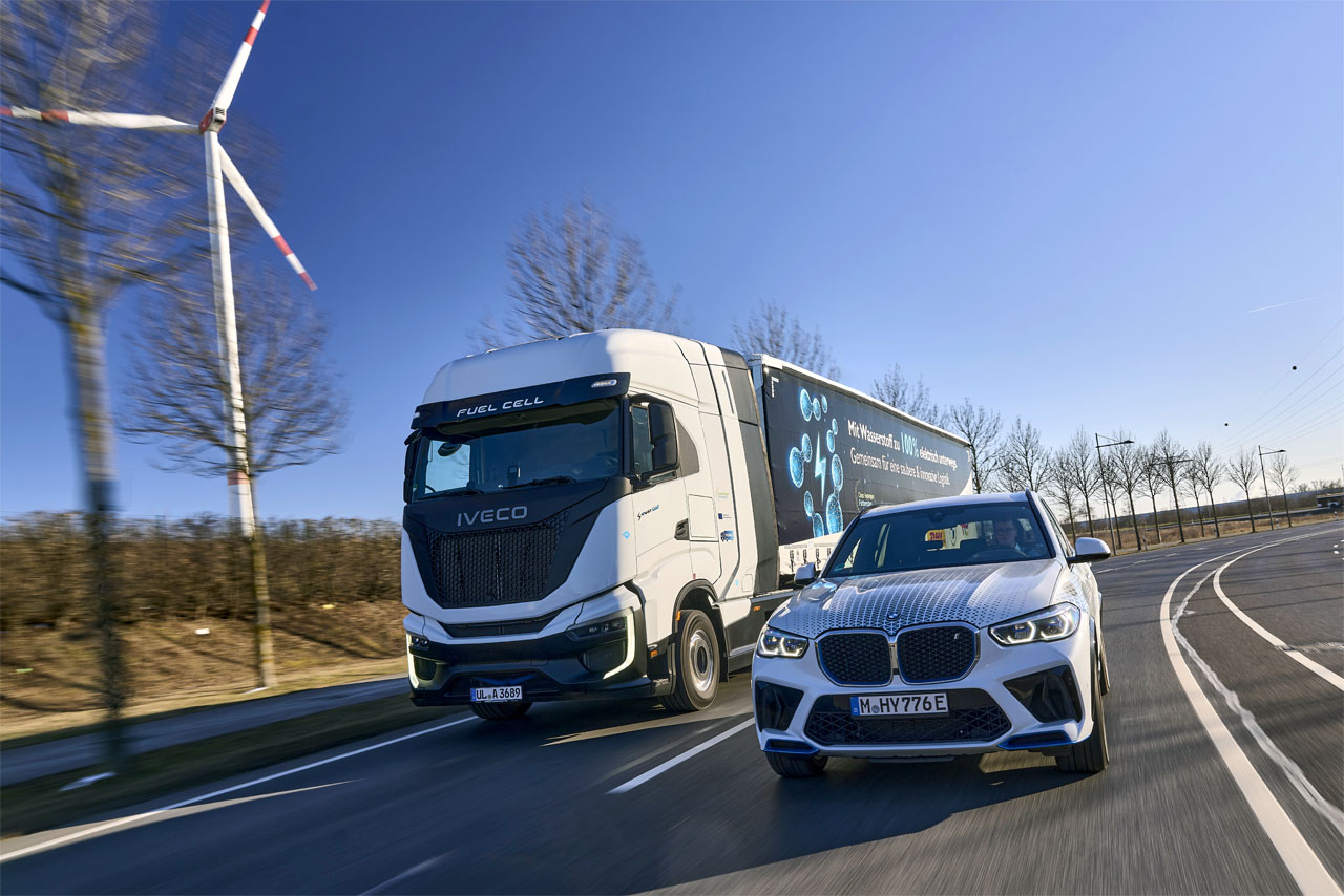 A BMW iX5 Hydrogen and an IVECO S-eWay Fuel Cell semi-trailer truck pass a wind turbine side by side on a multi-lane road.