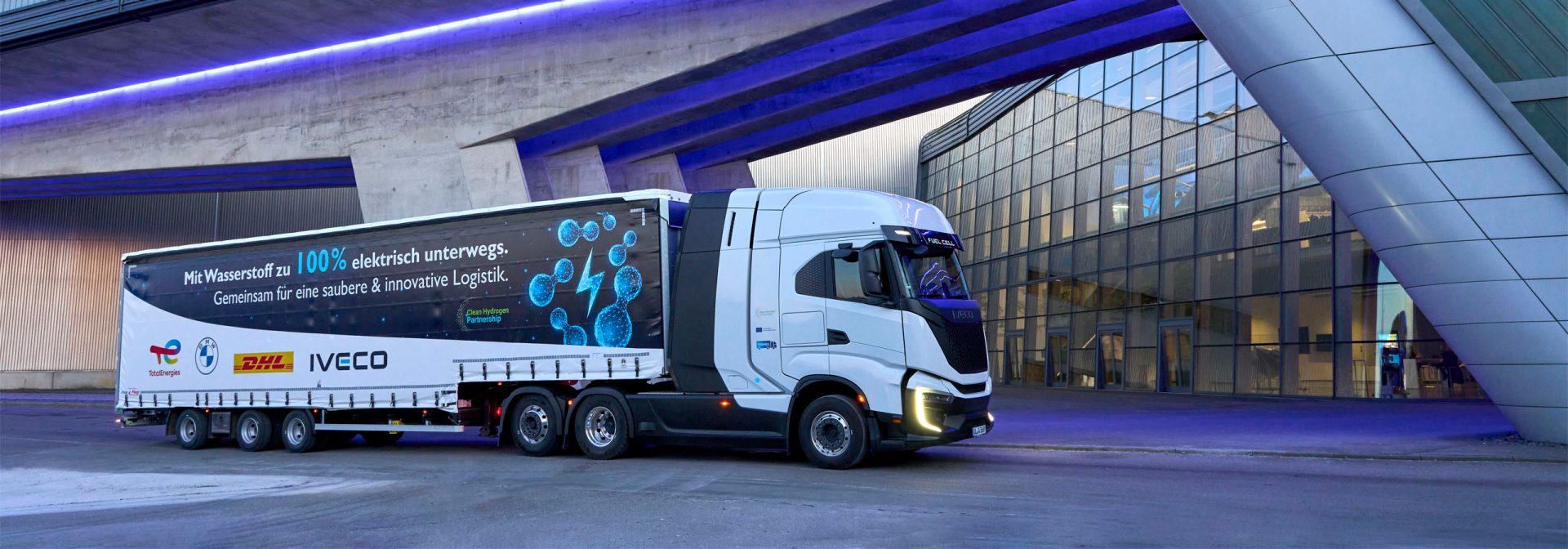 An IVECO S-eWay Fuel Cell semi-trailer truck in front of the illuminated, modern façade of BMW Group Plant Leipzig.