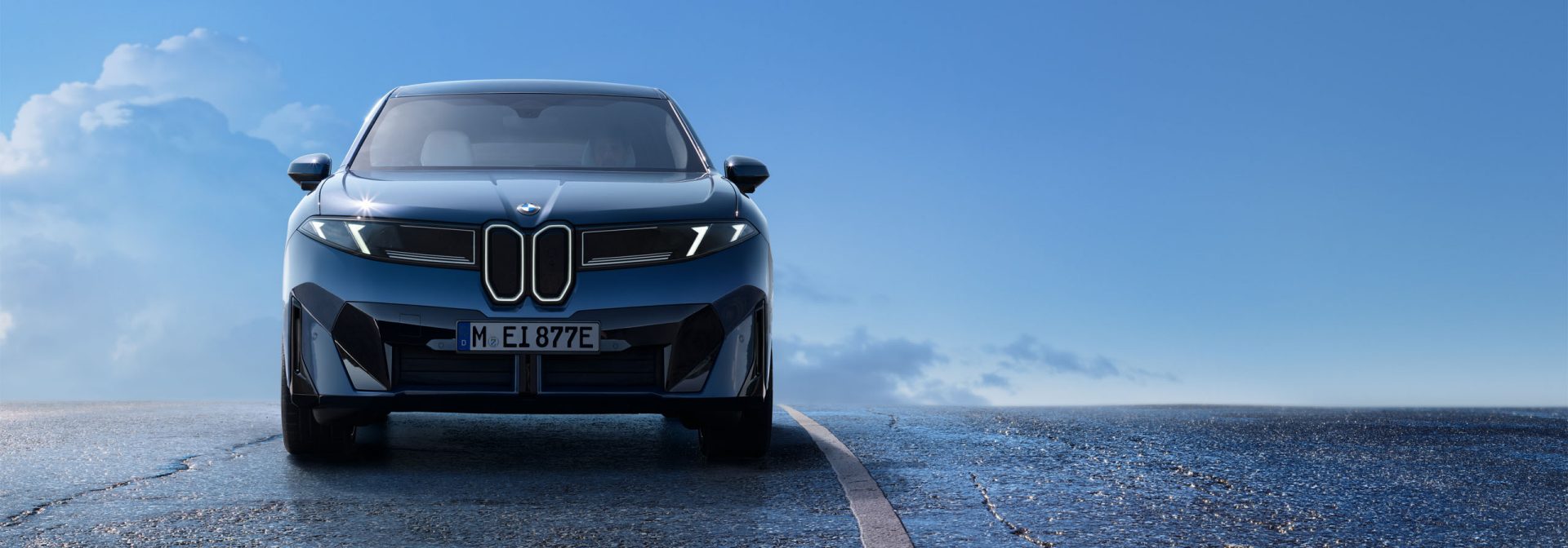 Front view of a dark-colored BMW iX3 on a road, with blue sky and clouds in the background.