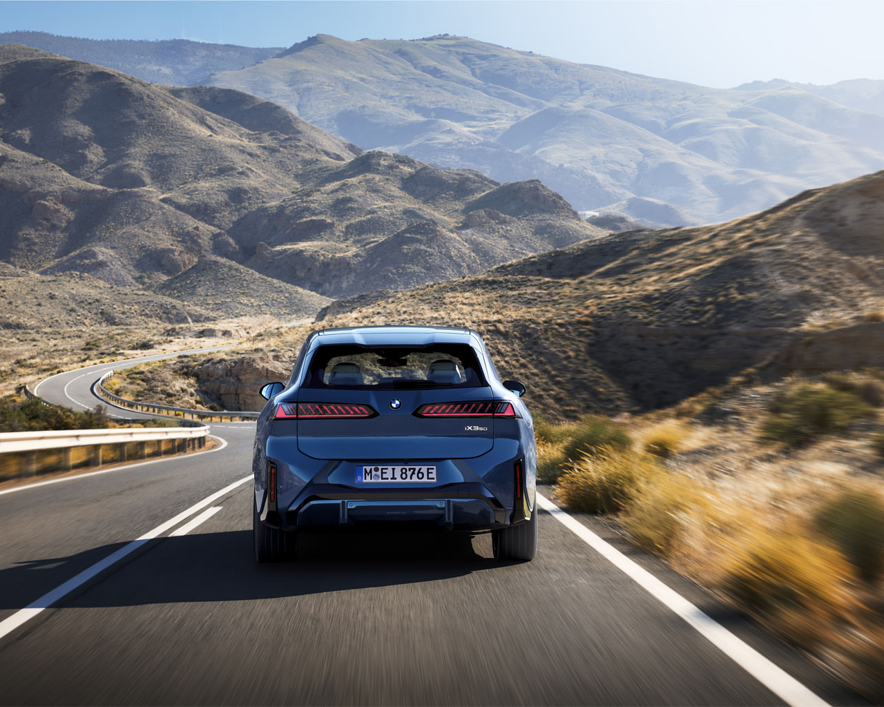 Rear view of a blue BMW iX3 driving on a winding road through a mountainous landscape with dry hills in the background.