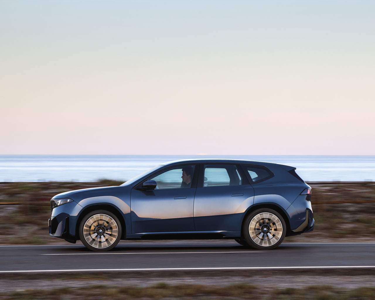 The new BMW iX3, a blue SUV, drives along a coastal road with the sea in the background.