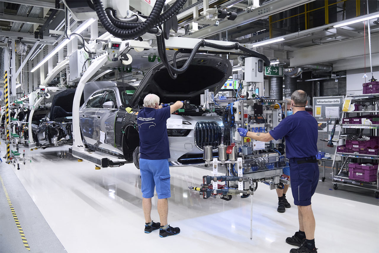 Assembly at BMW Group Plant Dingolfing: A BMW i7 hangs from a conveyor, its bonnet open, as three workers fit components.