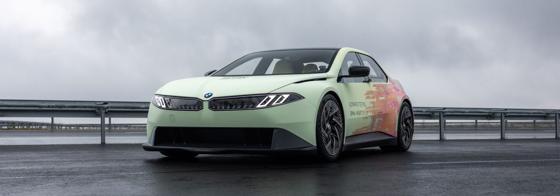 The BMW Vision Driving Experience viewed diagonally from the front at the edge of a test track with a crash barrier in the background.