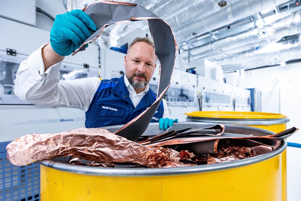 A BMW Group recycling specialist holds a black, foil-like material with copper-coloured edges in his hand to inspect it. The foil is stored in a yellow bin.