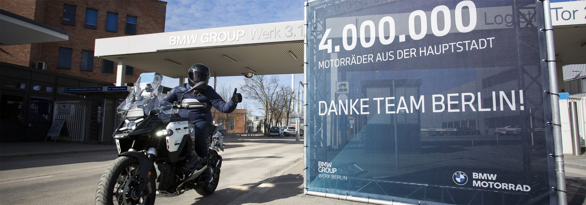 A motorcyclist leaves the premises of BMW Group Plant Berlin on a BMW R 1300 GS Adventure made at the facility. On the right, a large sign reads “4,000,000 motorcycles made in the capital – Thank you, Team Berlin!”