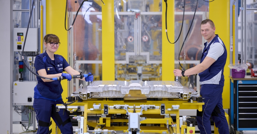 Two people are standing in a factory hall to the left and right of a construction part that they are working on with tools.