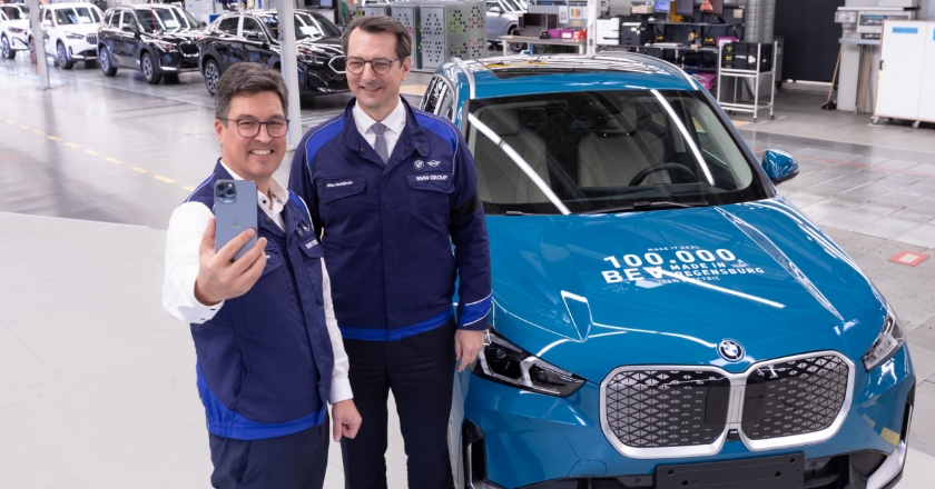 Two people stand in front of a BMW vehicle in a factory hall and take a selfie.