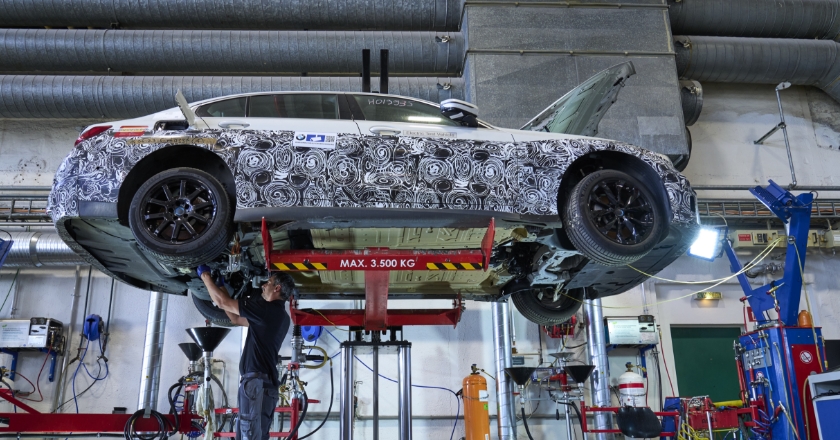 A BMW with camouflage film is standing on a lifting platform. A man is working on the underside of the car.