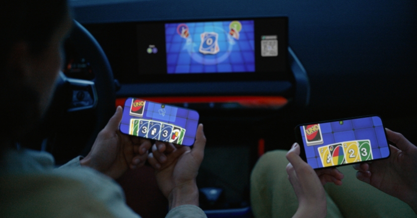 Two people are sitting in a vehicle with mobile phones in their hands. The game UNO® can be seen on the mobile phones and on the display in the cockpit.