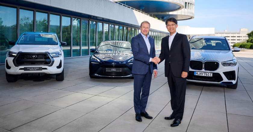 Two men stand in front of the BMW headquarters in Munich and shake hands. Behind them are three BMWs.