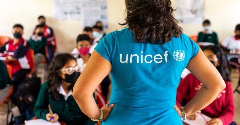 The “Unicef” logo is depicted on the back of a woman's T-shirt. She is teaching a school class.