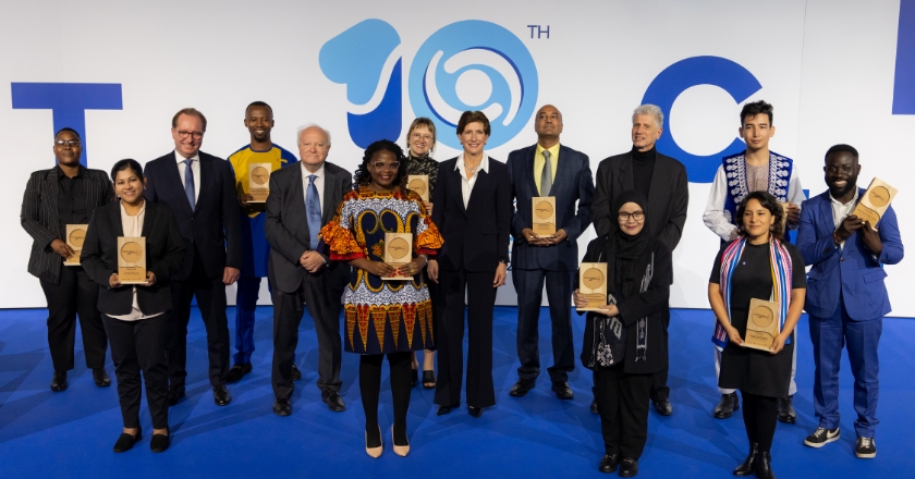 14 people from different nations stand in front of a wall with the number ten.