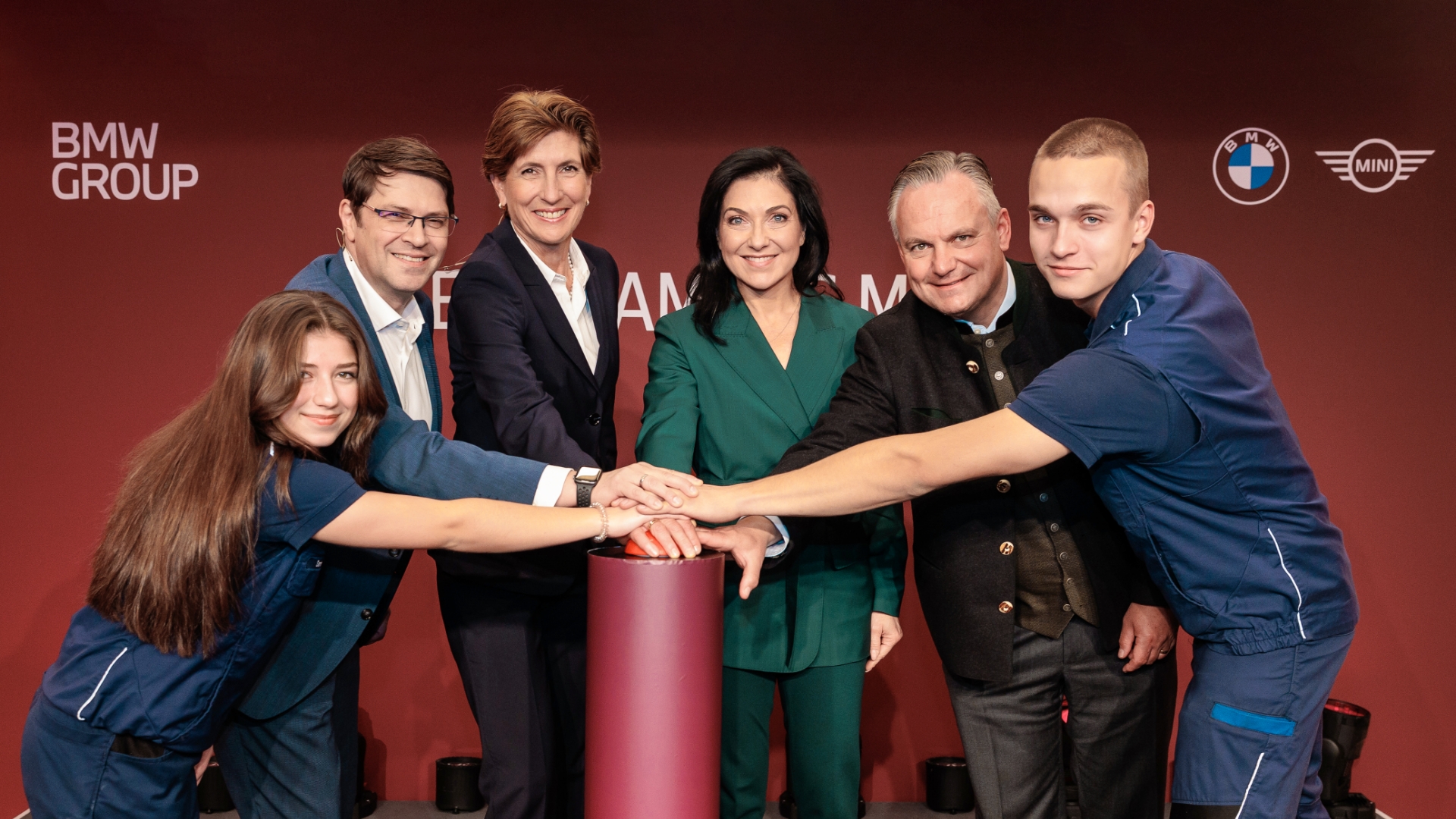 Group of different people placing their hands together on a large red button in front of a BMW Group backdrop.