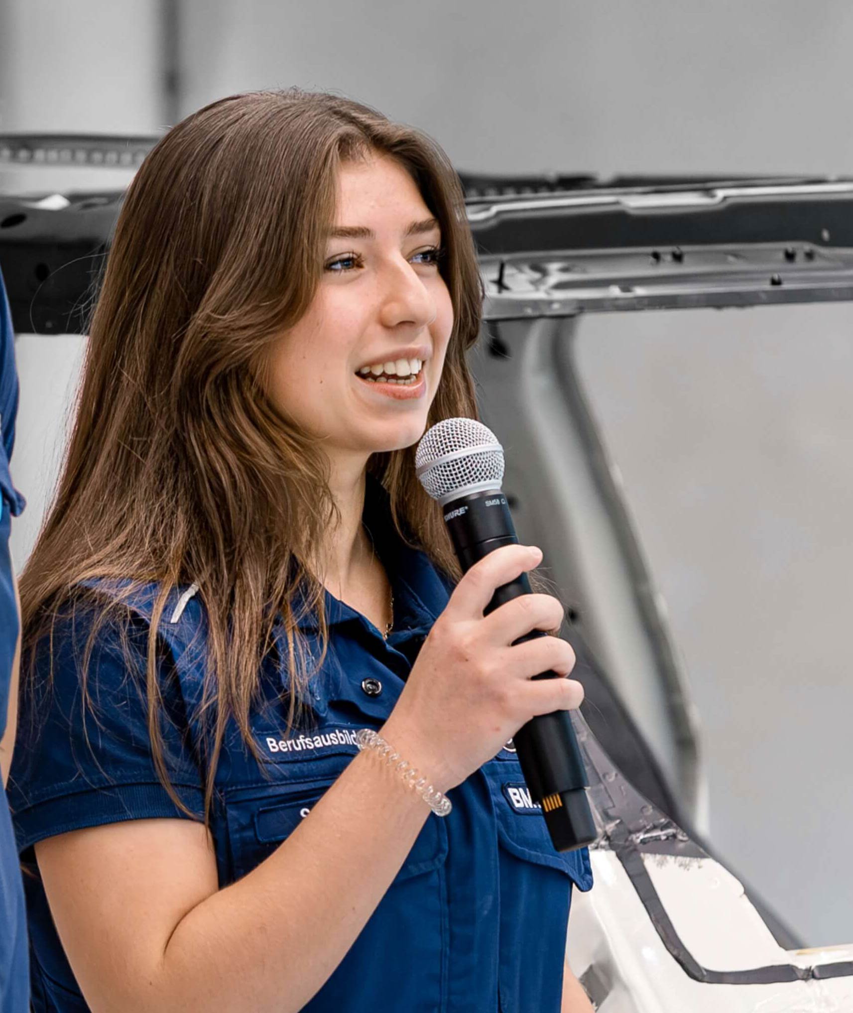 Two people in BMW workwear presenting in front of a vehicle body, one person speaking into a microphone.
