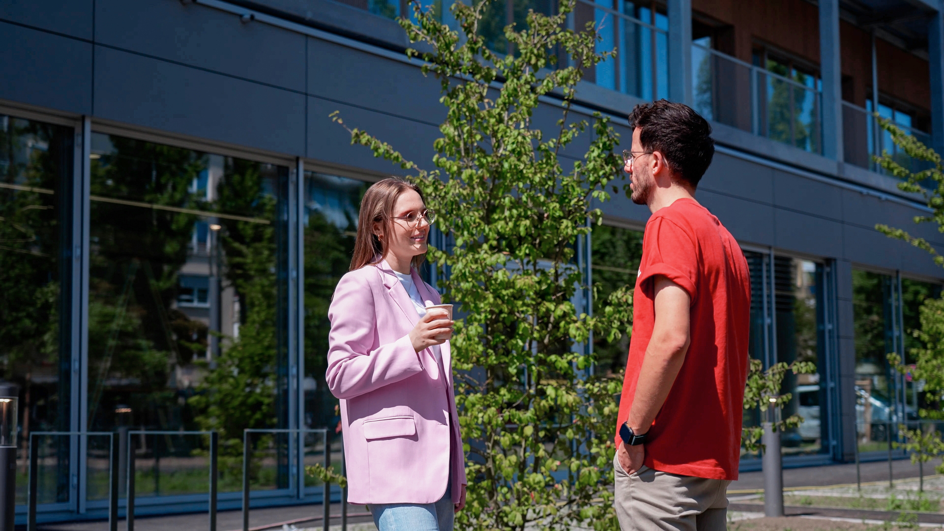 Two people talking outdoors in front of a modern building, one person holding a cup.