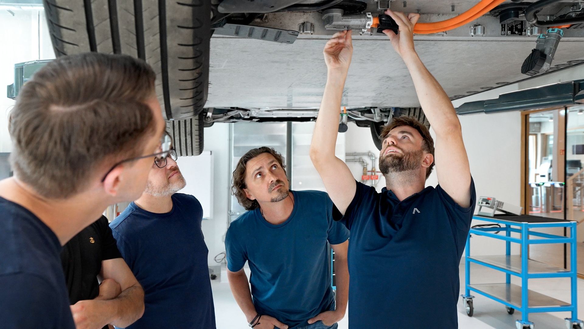 Group of people inspecting a lifted vehicle in a room, with one person working on components in the underbody.
