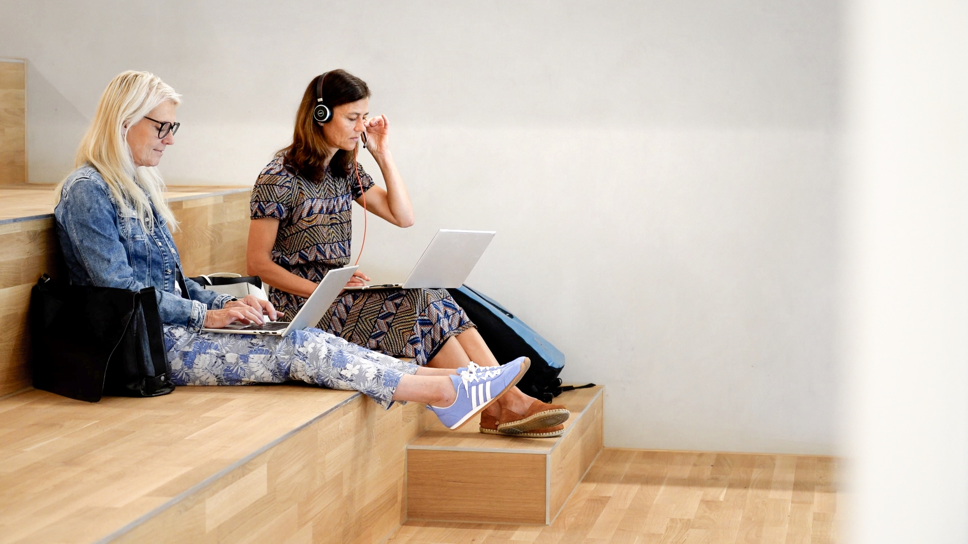 Two people working on laptops on wooden tiered seating in a modern interior, one person wearing headphones.