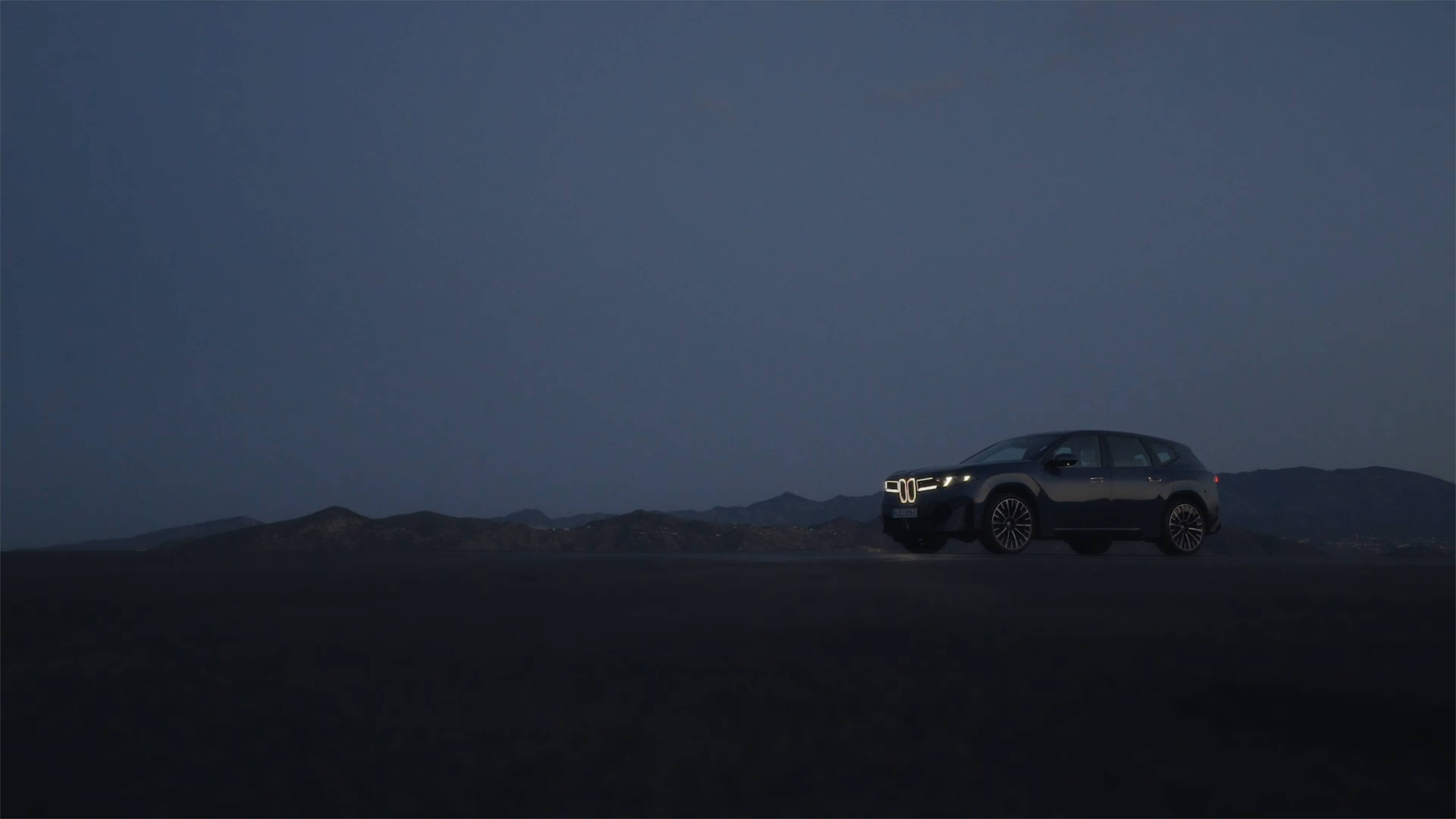 BMW vehicle with illuminated front in an open landscape at dusk, with silhouetted mountains in the background.