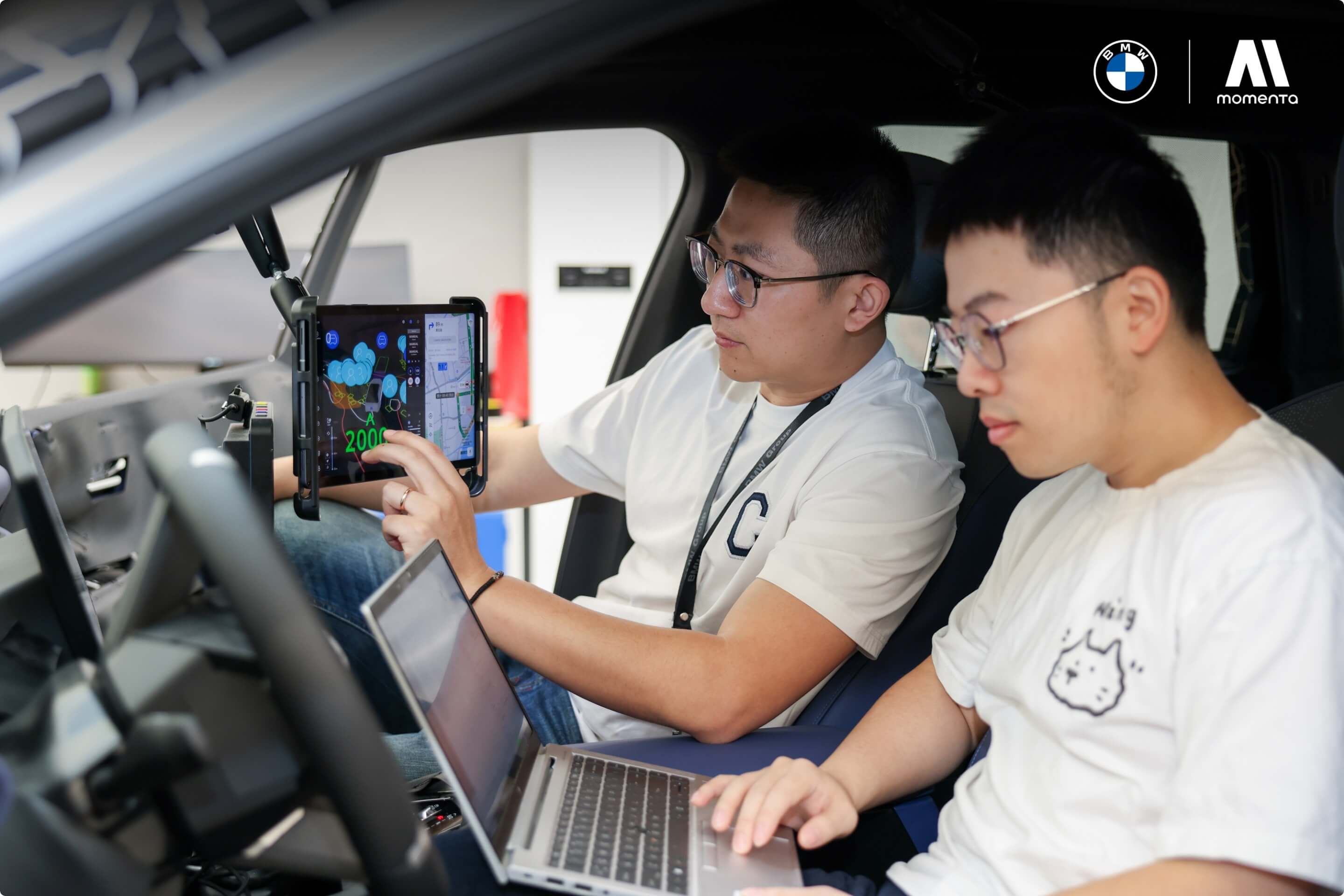 Two individuals work in a vehicle with tablet and laptop; BMW and MOMENTA logos are included in the image.