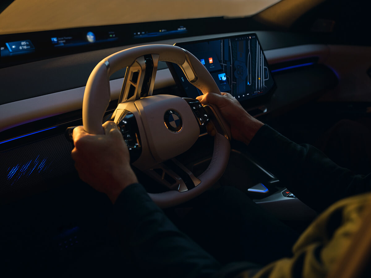 Driver holding a two‑spoke BMW steering wheel in front of a digital display in a dimly lit setting.