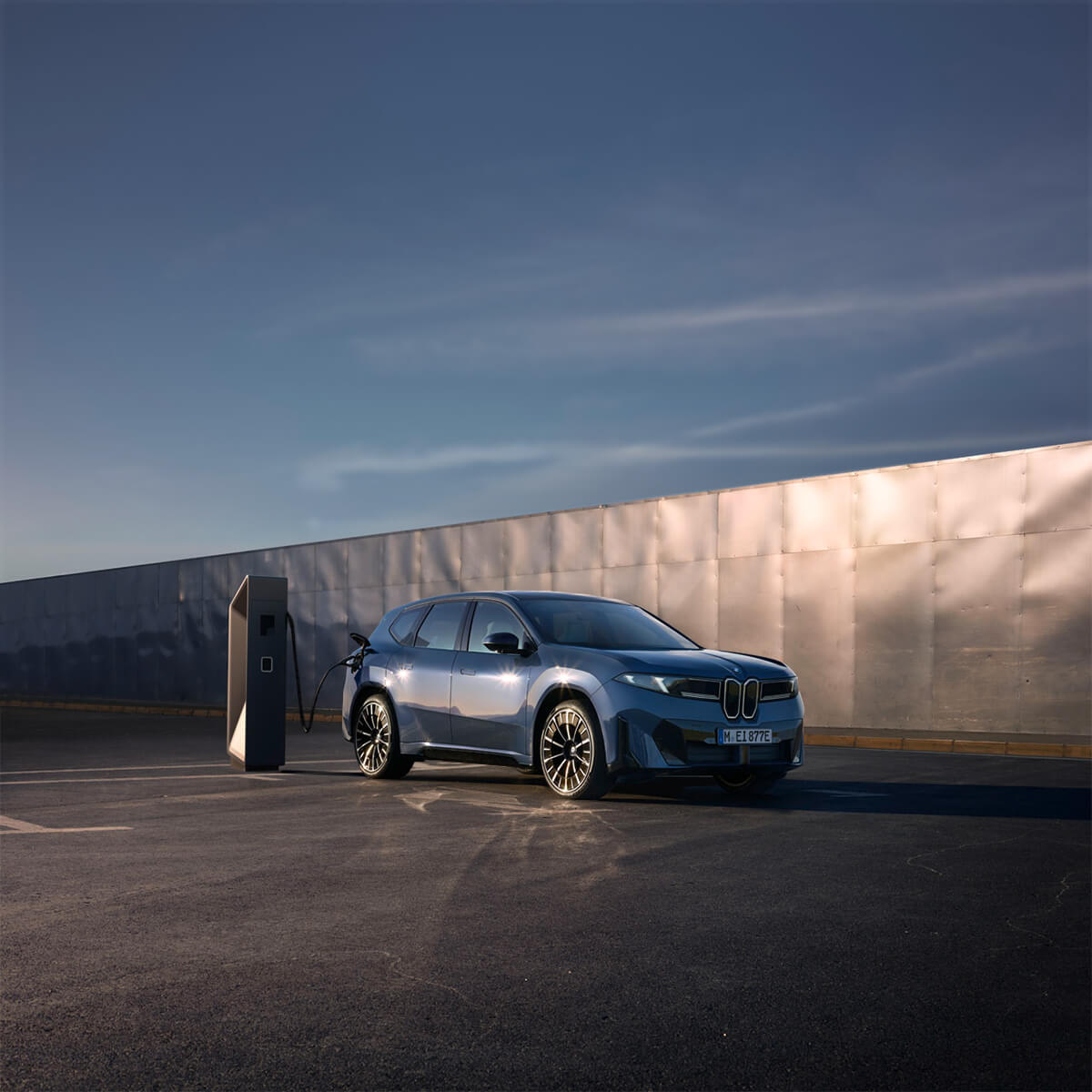BMW electric vehicle at a charging station on an open paved area in front of a metallic wall under a clear sky.