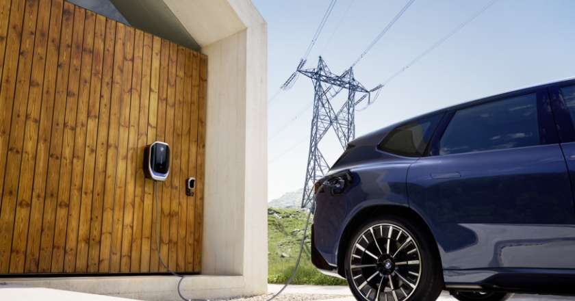 Electric vehicle is connected to a wallbox on a wooden facade, with high‑voltage power lines running across a rural landscape in the background.