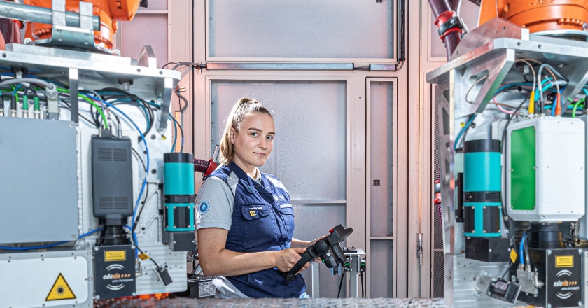 Employee stands between two automated manufacturing units operating a control device in a technical production environment.