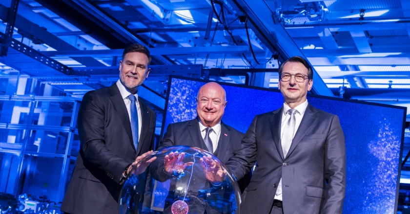 Three people in suits stand in a blue‑lit setting and jointly touch an illuminated glass sphere placed on a pedestal.