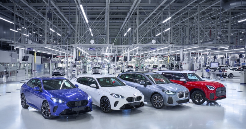 Four BMW vehicles of different models are positioned side by side in a bright production hall between several assembly lines.