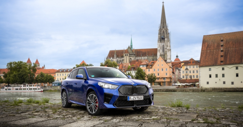 Blaues BMW‑Fahrzeug steht am Flussufer vor historischer Stadtkulisse mit markantem Kirchturm und Uferbebauung.