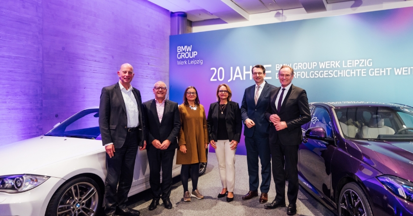 Six people stand between two BMW vehicles in front of a wall referencing “20 years of BMW Group Plant Leipzig.”