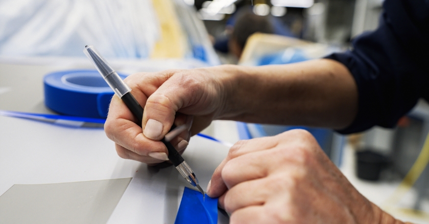 Hands use a precision knife to trim blue foil on a vehicle surface for a paint application process.
