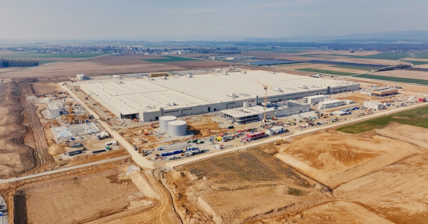Aerial view of a large BMW plant construction site with halls, cranes and construction vehicles, surrounded by fields and access roads.