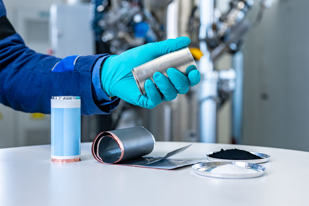 A person wearing a lab coat and turquoise gloves is holding a cylindrical metal object over a table.
