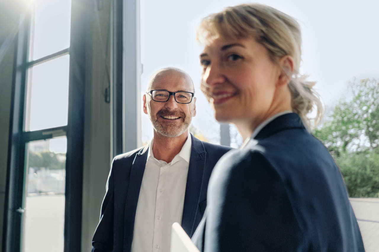 A man and a woman stand side by side in front of a large window overlooking the surrounding area.