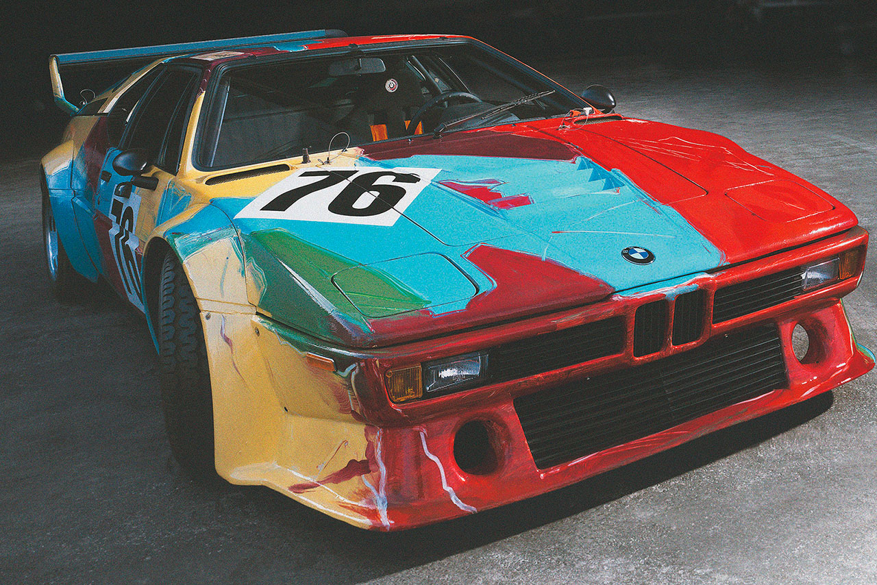 Colorful BMW M1 art car with number 76 displayed in a garage.